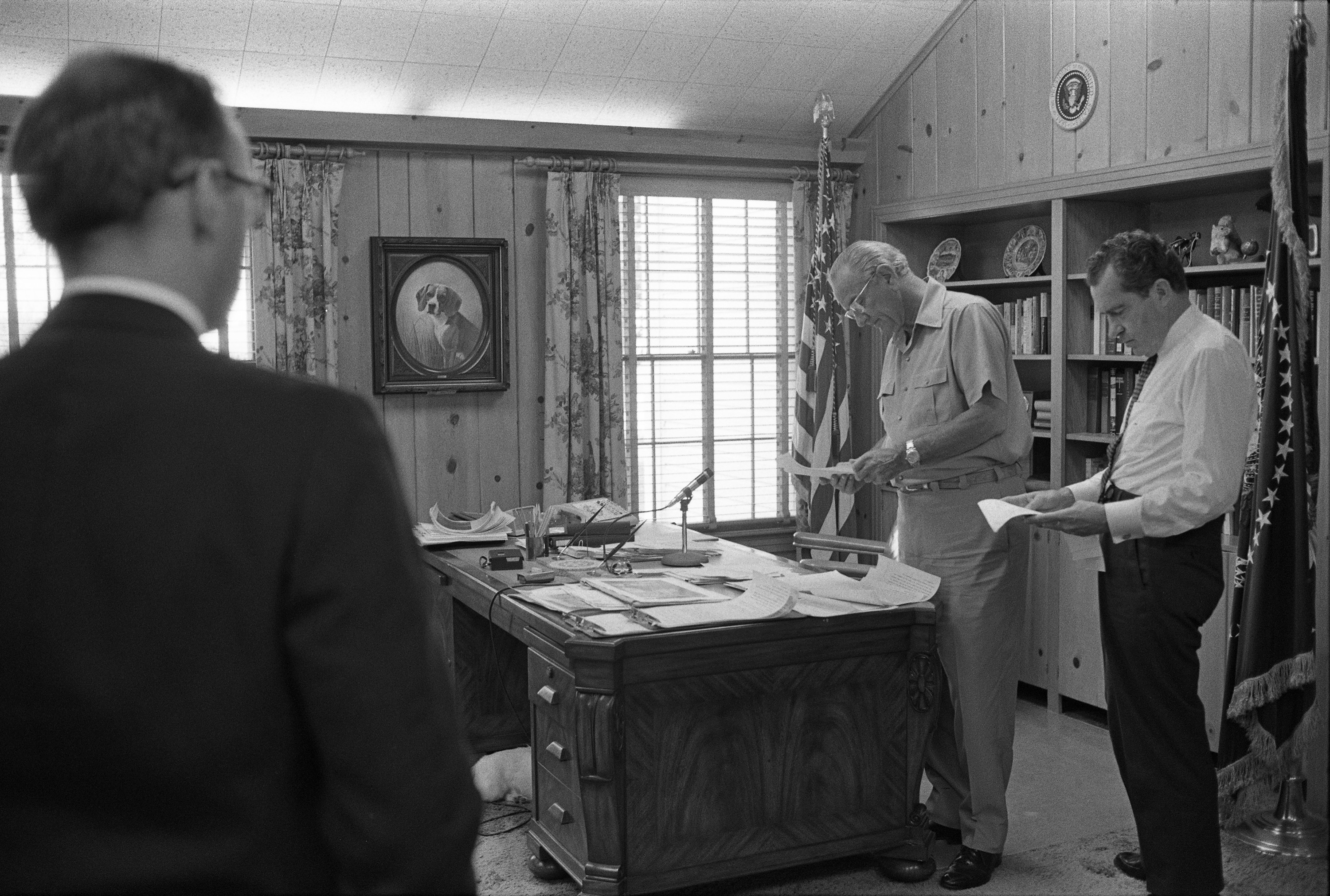On the left, a man stands with his back to the camera. LBJ and Richard Nixon stand behind a desk and look at papers. LBJ wears leisurewear. Nixon is in shirtsleeves with tie.